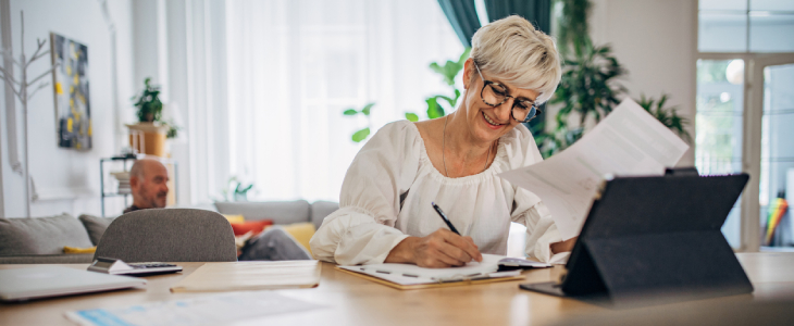 older woman with thick glasses smiling writing on paper with a tablet in front of her ssdb
